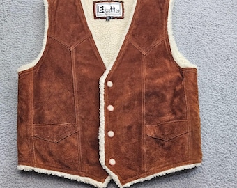 Könnte beinhalten: Eine braune Wildlederweste mit cremefarbenem Kunstfellfutter, V-Ausschnitt, vier Knöpfen auf der Vorderseite und zwei unteren Taschen. Das Innenetikett zeigt den Text Excelled. Die Weste hat eine strukturierte Oberfläche und sichtbare Nähte.