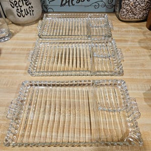 May include: Three rectangular clear glass serving trays with a ribbed texture and a scalloped edge. Each tray features a small, separate compartment on one side and two small handles on the ends, designed for serving food items.