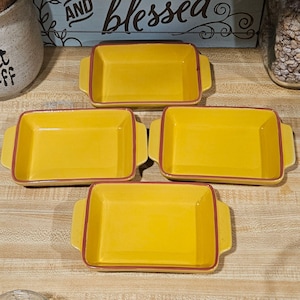 May include: Four rectangular yellow ceramic baking dishes with red trim and handles. The dishes are empty and arranged on a light-colored surface. The background includes a decorative sign that says "AND Blessed".