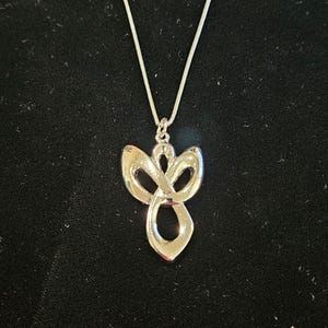 K&ouml;nnte beinhalten: Eine silberfarbene Metall-Halskette mit einem Anh&auml;nger in Form einer abstrakten, stilisierten Engelsfigur mit glatten, flie&szlig;enden Linien und einem durchbrochenen Design. Der Anh&auml;nger h&auml;ngt an einer d&uuml;nnen, passenden silberfarbenen Schlangenkette.