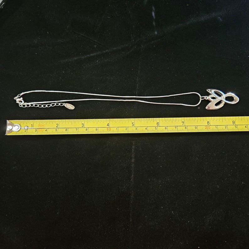 K&ouml;nnte beinhalten: Eine silberfarbene Halskette mit einem polierten, abstrakten Anh&auml;nger in geschwungenem Design. Die Kette verf&uuml;gt &uuml;ber ein verstellbares Verl&auml;ngerungsst&uuml;ck mit einem kleinen ovalen Anh&auml;nger. Ein gelbes Ma&szlig;band zeigt eine L&auml;nge von ca. 23 cm.