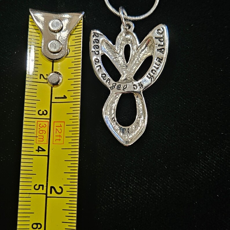 K&ouml;nnte beinhalten: Ein silberfarbener Metall-Engel-Anh&auml;nger an einer Kette, platziert neben einem gelben Ma&szlig;band, das eine L&auml;nge von etwa 5 cm anzeigt. Der Anh&auml;nger hat ein durchbrochenes Design mit dem eingravierten Text 'keep an angel by your side'.