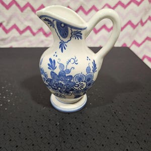 May include: A small, white ceramic jug with a handle and spout. The jug is decorated with blue floral patterns and designs. The base is round and has a blue trim. The jug is likely used for serving liquids.