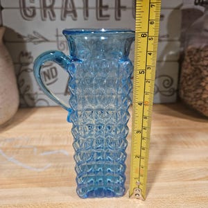 May include: A light blue glass pitcher with a handle and a textured diamond pattern. The pitcher is approximately 6 inches tall, as measured by a yellow ruler. The pitcher is likely vintage and suitable for serving beverages.