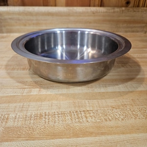 May include: A stainless steel bowl with a wide, flat rim. The bowl is round and has a shiny, reflective surface. The bowl is sitting on a wooden surface.