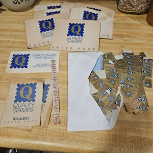 May include: A collection of vintage Quality Stamps saver books and a white envelope filled with loose brown and blue postage-style stamps. The books feature a blue square logo with a large letter Q and the text Quality Stamps Saver Book.