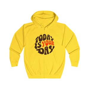 Peut inclure: Un sweat à capuche jaune avec fermeture éclair et poches avant. Le sweat-shirt affiche le texte "TODAY IS YOUR DAY" dans un design circulaire, avec des lettres marron, rouges et blanches. Le vêtement est en matière douce et confortable.