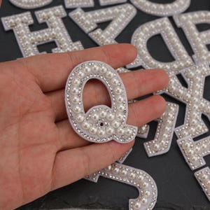 May include: White fabric letter "Q" embellished with faux pearls and rhinestones. The letter is surrounded by other letters with similar embellishments. The letters are on a dark surface.