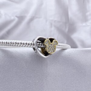 May include: A silver charm bracelet featuring a heart-shaped charm with a gold-toned Mickey Mouse head silhouette. The Mickey Mouse icon is encrusted with small, clear, sparkling stones, and the charm is threaded onto a braided silver chain.