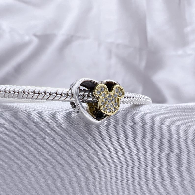 May include: A silver charm bracelet featuring a heart-shaped charm with a gold-toned Mickey Mouse head silhouette. The Mickey Mouse icon is encrusted with small, clear, sparkling stones, and the charm is threaded onto a braided silver chain.