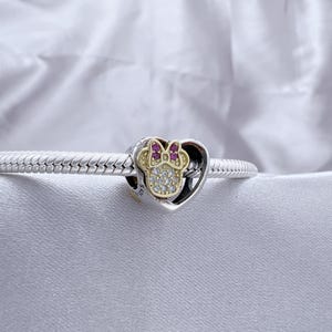 May include: A silver snake chain bracelet featuring a heart-shaped charm. The charm contains a gold-toned Minnie Mouse head silhouette adorned with clear cubic zirconia stones and a small bow accented with pink stones, marked with S925.
