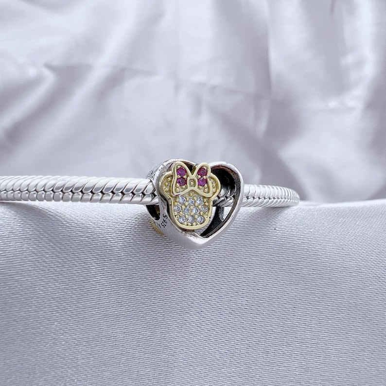 May include: A silver snake chain bracelet featuring a heart-shaped charm. The charm contains a gold-toned Minnie Mouse head silhouette adorned with clear cubic zirconia stones and a small bow accented with pink stones, marked with S925.