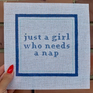 May include: A square white needlepoint canvas featuring a dark blue border and a light blue inner frame. The text "just a girl who needs a nap" is printed in dark blue in the center of the canvas. The design is on a grid-like mesh fabric.