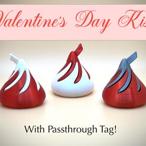 May include: Three decorative kiss-shaped objects in red, white, and blue, with a Valentine's Day theme. The objects have a textured appearance and are arranged on a light background. The text "Valentine's Day Kiss" is displayed above the objects.