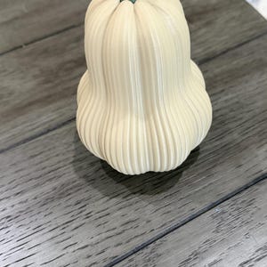 May include: A cream-coloured, vertically ribbed gourd-shaped decorative object with a smooth, matte finish. The object features a distinct, solid sage green stem at the top, which is shaped like a small, faceted hexagonal prism.