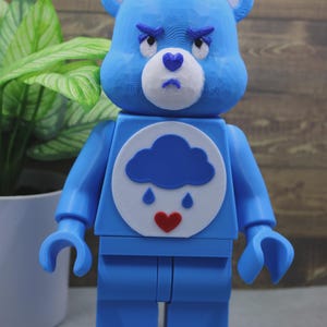 Pode incluir: Uma figura em forma de urso azul com um corpo articulado e quadrado. O rosto apresenta um focinho branco, um nariz azul em forma de coração e uma expressão carrancuda. O peito exibe um emblema circular branco com uma nuvem azul, duas gotas de chuva azuis e um coração vermelho.