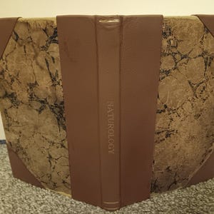 May include: A vintage book with a brown leather spine and corners. The book's covers feature a marbled pattern in shades of brown and tan. The word "NATUROLOGY" is printed vertically on the spine.