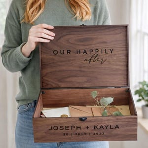 May include: A wooden keepsake box with the words "OUR HAPPILY after" engraved on the lid. The box is open, revealing envelopes and greenery inside. The bottom of the box is engraved with "JOSEPH + KAYLA 30 | JULY | 2023".