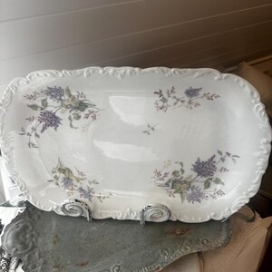 May include: A white rectangular serving platter with a scalloped edge, adorned with purple and green floral designs. The platter is supported by two decorative metal stands. A gray, decorative tray is beneath the platter.