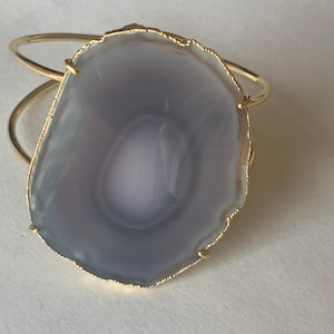 May include: A gold-toned cuff bracelet featuring a large, polished agate stone. The stone is primarily gray with lighter, concentric bands and a gold-colored edge. The bracelet has a simple, open design.