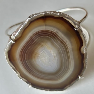 May include: A silver bracelet featuring a polished agate stone. The agate displays concentric bands of brown, tan, and white. The stone is mounted on a simple, elegant silver band, creating a natural and stylish accessory.