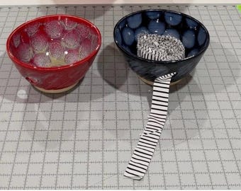 May include: Two ceramic bowls with a textured, crater-like interior glaze. The left bowl is red with white highlights, and the right bowl is dark blue with light blue highlights. A black and white striped measuring tape is inside the blue bowl.