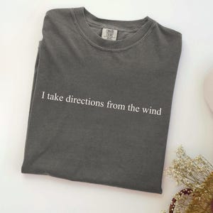 May include: Dark grey t-shirt with the text "I take directions from the wind" in white. The shirt has a crew neck and short sleeves. The Comfort Colors tag is visible at the neckline.