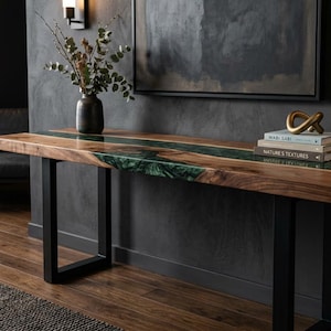 May include: A wooden console table with a live edge design and a green epoxy river. The table has black metal legs and is decorated with books, a gold sculpture, and a vase of greenery. The table is in a room with dark walls.
