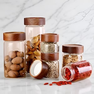 Op de afbeelding: Een verzameling heldere glazen potten met houten deksels, gevuld met verschillende noten, zaden en gedroogde bessen. De potten staan op een wit marmeren oppervlak, waarbij de inhoud wordt getoond.