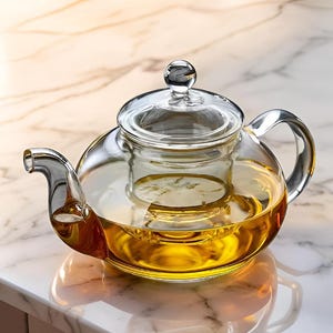 May include: A clear glass teapot with a glass infuser and lid. The teapot is filled with a golden-colored liquid, likely tea. The handle and spout are curved. The teapot sits on a marble surface.