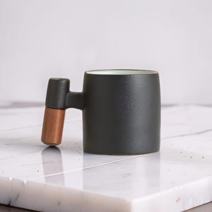 Peut inclure: Une tasse en céramique noire avec une poignée originale. La poignée est composée d'une partie sombre et rectangulaire reliée à une prise en bois. La tasse a une surface texturée et un intérieur clair. La tasse est posée sur une surface en marbre blanc.