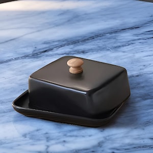 Peut inclure: Un beurrier en céramique noire avec un couvercle carré et un bouton en bois clair. Le plat repose sur une base rectangulaire assortie. Le beurrier est un article de cuisine.