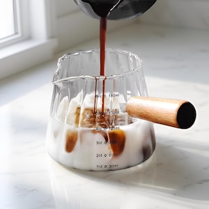 May include: A clear glass pitcher with a wooden handle is being filled with a dark liquid, likely coffee, over ice and a white liquid. The pitcher has measurement markings in ounces and milliliters.