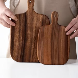 Peut inclure: Deux planches à découper en bois avec une poignée et un trou pour suspendre. Les planches sont en bois brun foncé avec des motifs de grain visibles. Les planches sont rectangulaires avec des coins arrondis.