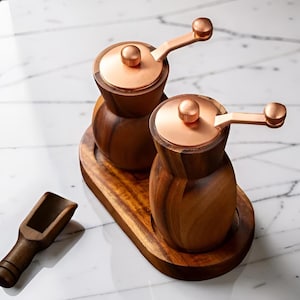 Peut inclure: Un ensemble de moulins à sel et à poivre en bois avec des couvercles et des poignées cuivrées, présenté sur un plateau en bois. Une petite cuillère en bois est également incluse, le tout sur une surface en marbre. Les moulins sont en bois foncé.