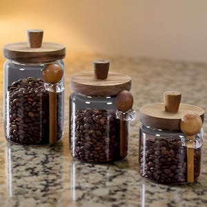 Puede incluir: Tres frascos de vidrio llenos de granos de café, cada uno con una tapa de madera y un pequeño pomo de madera en la parte superior. Cada frasco tiene una cuchara de madera con un mango transparente. Los frascos están sobre una encimera moteada.