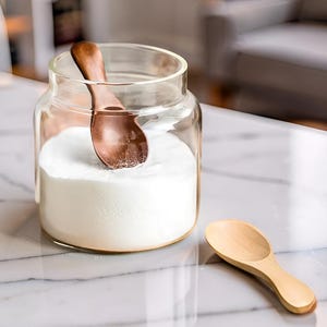 Puede incluir: Un tarro de cristal transparente lleno de polvo blanco, con una cuchara de madera dentro. Una segunda cuchara de madera descansa sobre la superficie. Las cucharas son de color marrón claro. El tarro está sobre una superficie de mármol blanco.