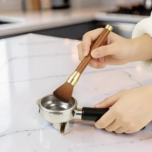 Peut inclure: Un porte-filtre de machine à café est nettoyé avec une brosse à poils bruns avec un manche en bois et des accents dorés. Le porte-filtre est en acier inoxydable avec une poignée noire. La brosse est tenue au-dessus du porte-filtre.