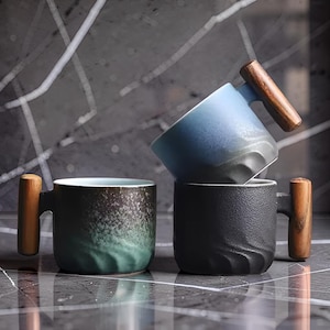 Peut inclure: Trois tasses en céramique avec des poignées en bois. L'une est noire, une autre est verte et noire, et la troisième est bleue et noire. Les tasses sont empilées les unes sur les autres. Les tasses ont un design texturé.