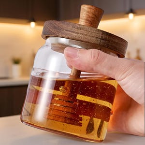 Könnte beinhalten: Ein klares Honigglas mit einem Holzdeckel und einem Dipper. Das Glas ist mit goldenem Honig gefüllt. Der Holzdeckel und der Dipper haben eine natürliche Holzmaserung. Das Glas wird von einer Hand gehalten.