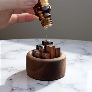 Peut inclure: Diffuseur d'huiles essentielles en bois avec un design géométrique. Une bouteille en verre marron verse de l'huile sur les blocs de bois. Le diffuseur est en bois brun foncé et repose sur une surface en marbre blanc.