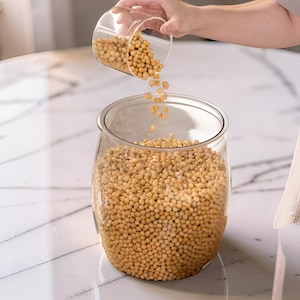 Peut inclure: Un bocal en verre transparent rempli de graines de soja jaunes. Un autre récipient en verre transparent est utilisé pour verser plus de graines de soja dans le bocal. Le bocal est posé sur une surface en marbre blanc.