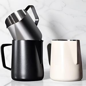 May include: Three milk frothing jugs in a stack. The top jug is stainless steel, the middle one is black, and the bottom one is off-white. Each jug has a handle and a spout for pouring. The jugs are on a white marble surface.