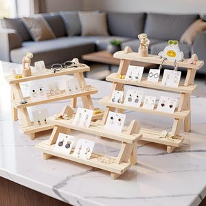 Può includere: Tre piccoli espositori in legno di colore chiaro, ciascuno con tre livelli, sono disposti su una superficie bianca. Gli espositori sono riempiti con varie paia di orecchini su cartoncini bianchi. Figurini decorativi sono posti sopra.