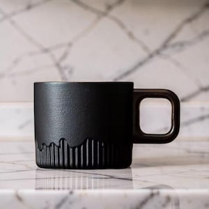 Peut inclure: Une tasse en céramique noire mate avec une surface texturée et une poignée carrée. Le bas de la tasse présente un motif rainuré vertical. La tasse est posée sur une surface en marbre.