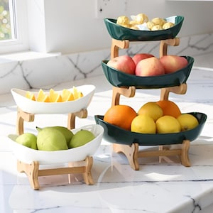 Op de afbeelding: Een fruitstandaard met meerdere niveaus met drie niveaus, elk met verschillende soorten fruit. De bovenste laag heeft bloemkool, de middelste laag rode appels en de onderste laag sinaasappels en citroenen. De stands zijn donkergroene en witte keramische kommen met houten steunen.