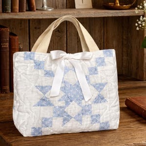 May include: A quilted tote bag featuring a white and light blue floral star pattern. The bag has two cream-colored fabric handles and a white satin ribbon tied into a bow at the center top. The quilted fabric has a diamond-stitched texture.