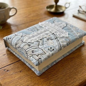 May include: A rectangular book cover made of quilted fabric featuring a light blue and white paisley pattern. A white lace cross is stitched onto the center of the cover. The fabric is padded and textured, providing a soft exterior for the book.