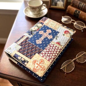 May include: A rectangular book cover featuring a patchwork design of various fabric patterns, including a central dark blue panel with white polka dots and a prominent light pink cross motif, surrounded by floral, geometric, and ornate textile prints.