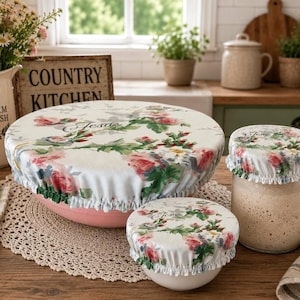 May include: Three elasticized fabric bowl covers featuring a white background with a floral pattern of pink roses, green leaves, and the word Jesus printed in script. The covers are shown fitted over a large pink bowl, a small white bowl, and a glass jar.